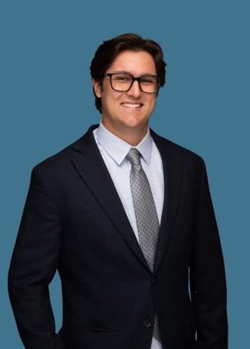 Smiling man wearing glasses, a dark suit and patterned tie against a gray-blue background.