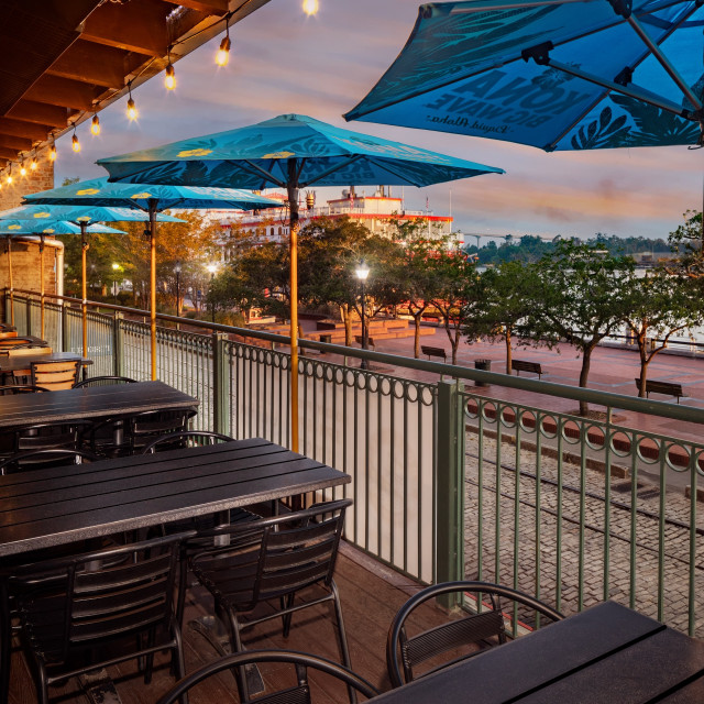 Outdoor balcony dining tables with blue umbrellas overlooking a river.