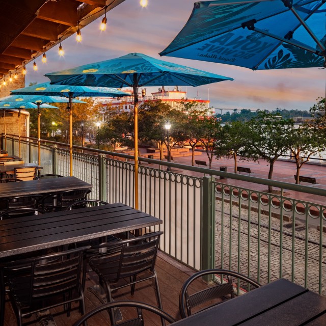Outdoor balcony dining tables with blue umbrellas overlooking a river.