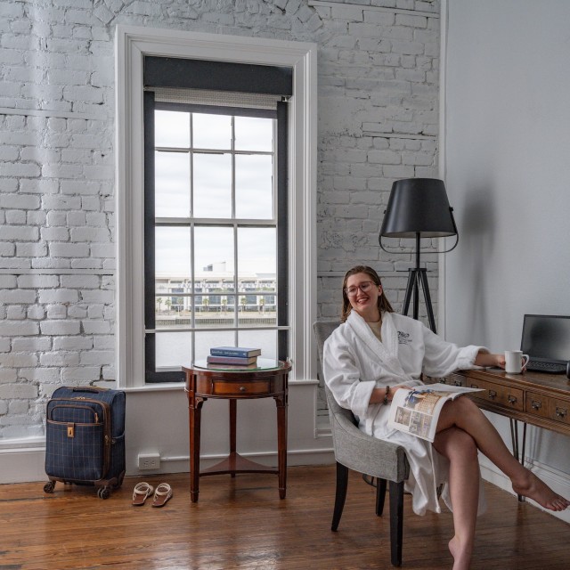 A young woman lounging in a chair in a hotel room wearing a white bathrobe.