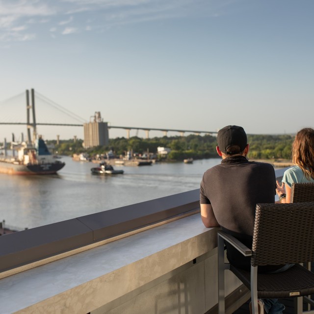 A man and woman overlooking the Savannah River on a rooftop deck as ships go by.