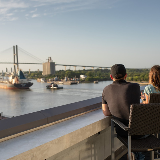 A man and woman overlooking the Savannah River on a rooftop deck as ships go by.