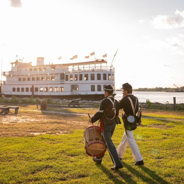 Two men dressed as soldiers during reenactment walking beside a riverboat.