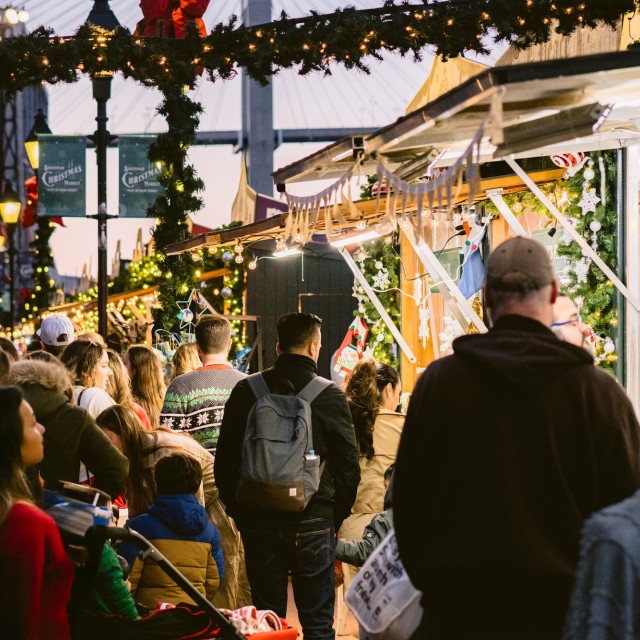 Crowds gather under garlands and twinkling lights at the Savannah Christmas Market at Plant Riverside District, exploring festive vendor stalls decorated for the holidays.