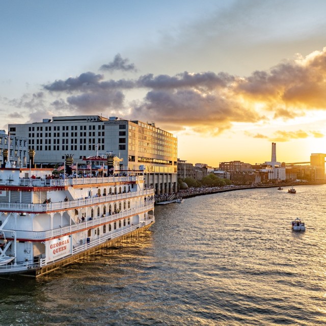 The Georgia Queen riverboat cruises along the Savannah River at sunset, with the city skyline and Talmadge Memorial Bridge glowing in the background.