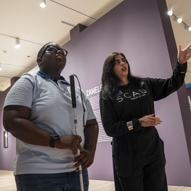 A visitor who is blind tours the SCAD Museum of Art with a guide describing an exhibit in the gallery.