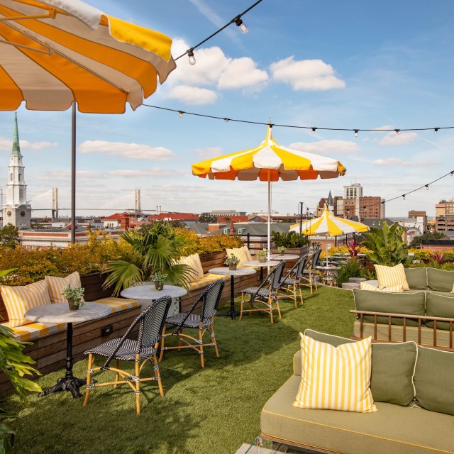 Outdoor rooftop lounge at Perry Lane Hotel’s Peregrin Rooftop Bar, featuring green seating, yellow-and-white umbrellas, café tables and sweeping views of the Savannah skyline and church steeple under a blue sky.