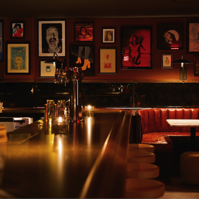 Dimly lit speakeasy-style lounge with red leather banquettes, candlelit tables, framed art-lined walls, and an intimate bar setting.