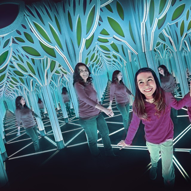 Girl exploring a mirrored light tunnel exhibit with geometric LED flooring and tree-shaped panels.