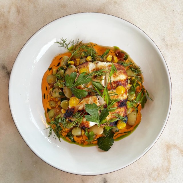 Seared fish with seasonal vegetables and herbs in a white bowl.