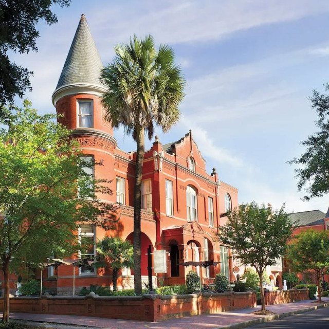 Exterior of Hotel Bardo Savannah, a red-brick historic building with a turreted corner, arched windows, palm trees and a tree-lined street in the foreground.