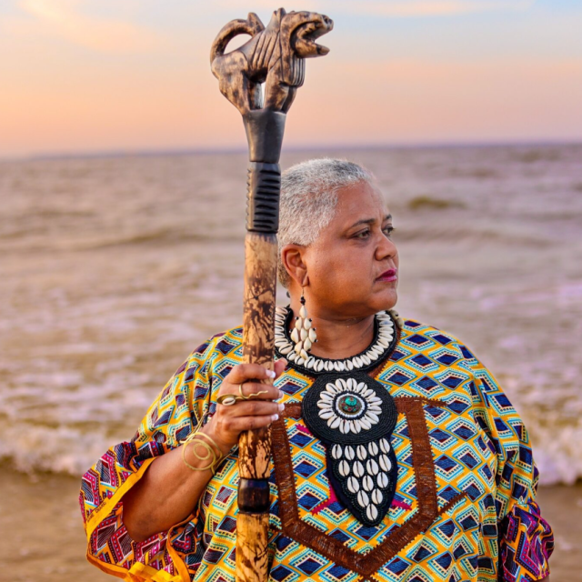 A woman dressed in African attire holding a staff while looking out onto the ocean.