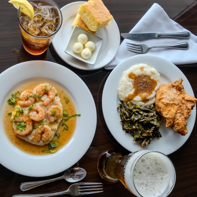 Southern-style meal with shrimp and grits, fried chicken, collard greens and drinks served at a dining table.