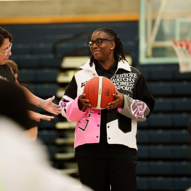 Woman holding a basketball on an indoor court, wearing a jacket that reads “Everyone Watches Women’s Sports,” with teammates nearby.