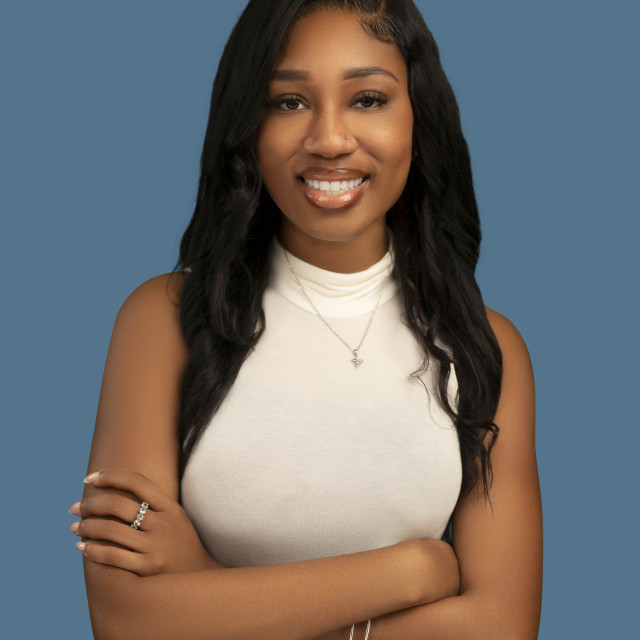 Smiling woman with long, wavy black hair and crossed arms, wearing a sleeveless white top against a blue background.