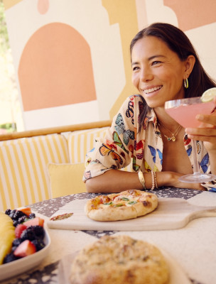 A woman smiles over a pink cocktail with pizza and fresh fruit on the table at a Savannah restaurant.