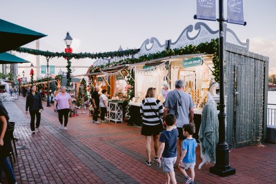 Visitors stroll through the Savannah Christmas Market, lined with festive vendor stalls decorated with garlands, bows, and twinkling lights.