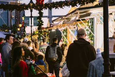 Crowds gather under garlands and twinkling lights at the Savannah Christmas Market at Plant Riverside District, exploring festive vendor stalls decorated for the holidays.
