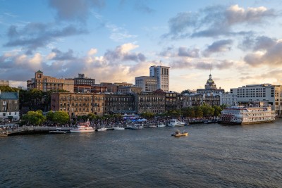 A view of Savannah’s historic River Street at sunset, with boats docked along the riverfront, crowds enjoying the waterfront, and the city skyline—including the gold-domed City Hall—glowing in the evening light.