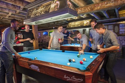 Group of friends playing pool inside a lively Savannah bar, gathered around a blue felt pool table with drinks in hand beneath exposed wooden beams.