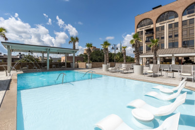 Outdoor pool deck at The DeSoto Savannah featuring a shallow sun shelf with in-water lounge chairs, palm trees, poolside seating and views of the hotel's rooftop terrace beneath a bright blue sky.