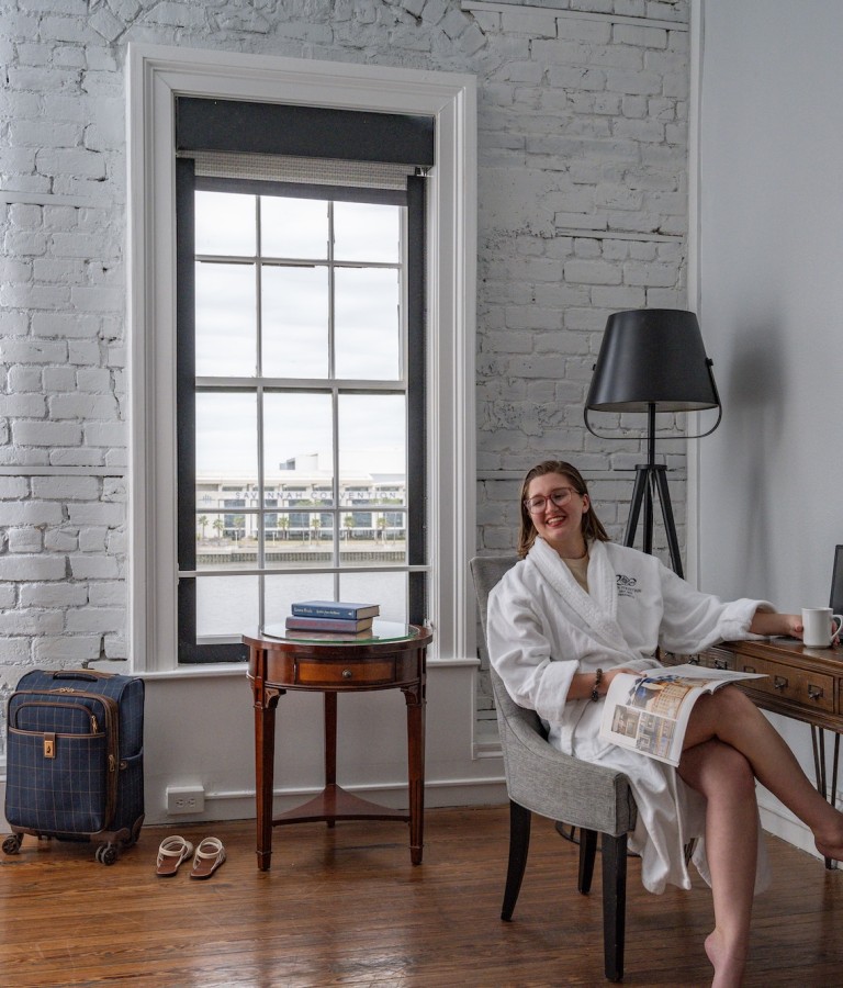 A young woman lounging in a chair in a hotel room wearing a white bathrobe.