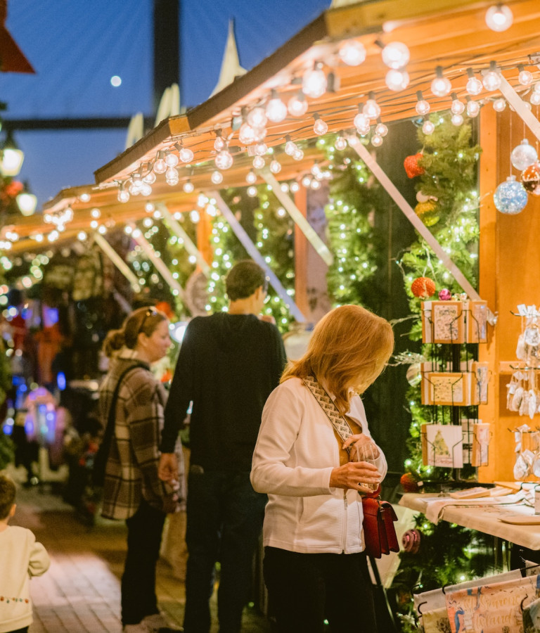 Shoppers browse festive stalls decorated with wreaths, ornaments, and twinkling lights at the Savannah Christmas Market in Plant Riverside District.