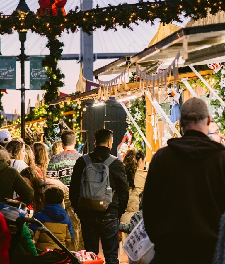 Crowds gather under garlands and twinkling lights at the Savannah Christmas Market at Plant Riverside District, exploring festive vendor stalls decorated for the holidays.