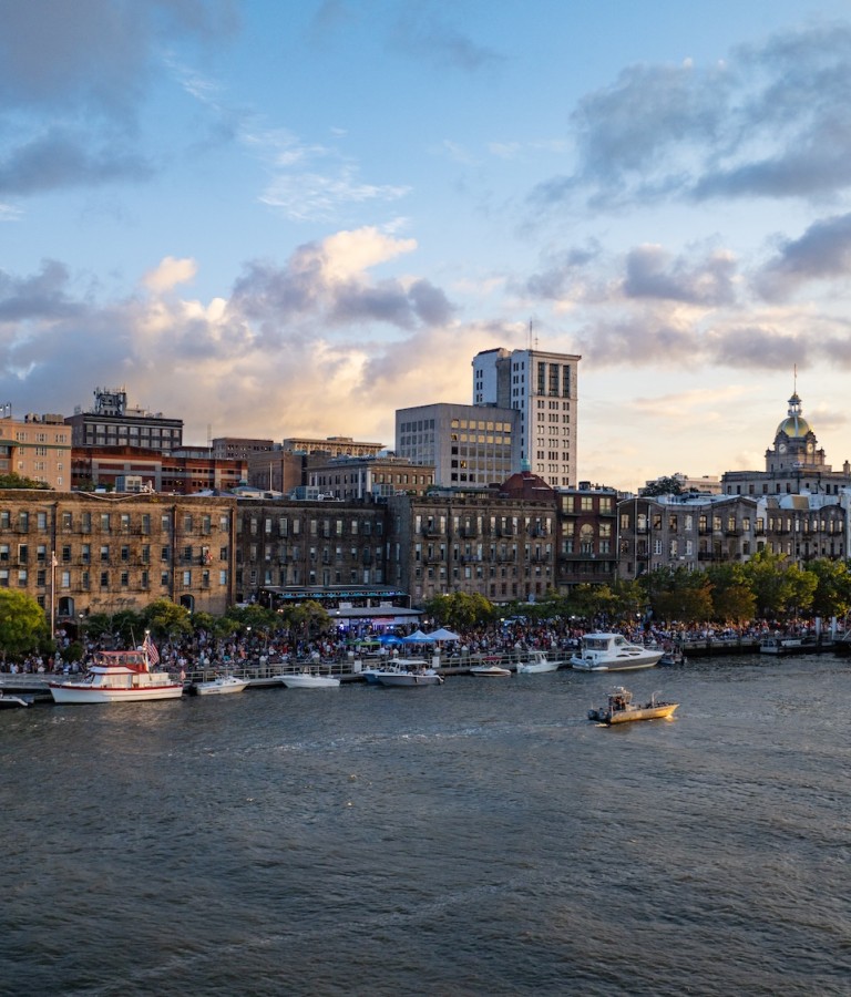 A view of Savannah’s historic River Street at sunset, with boats docked along the riverfront, crowds enjoying the waterfront, and the city skyline—including the gold-domed City Hall—glowing in the evening light.
