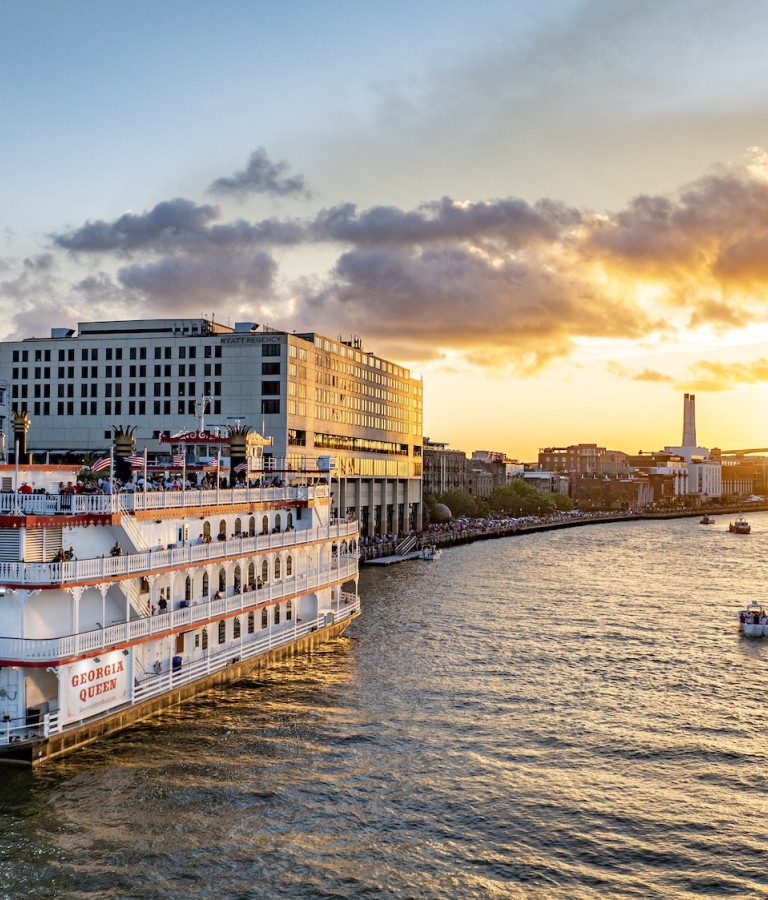 The Georgia Queen riverboat cruises along the Savannah River at sunset, with the city skyline and Talmadge Memorial Bridge glowing in the background.