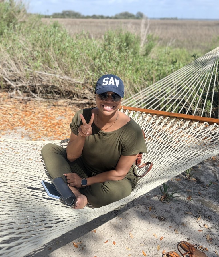 A woman wearing a baseball hat while sitting in a hammock on a beach.