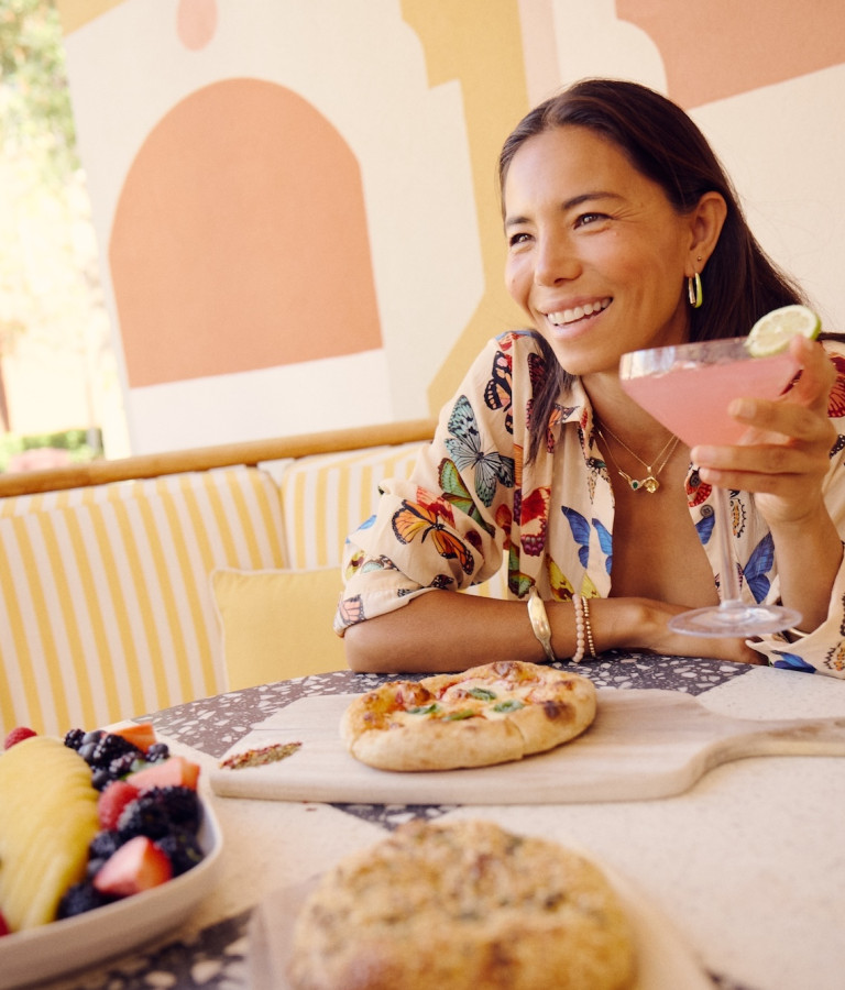 A woman smiles over a pink cocktail with pizza and fresh fruit on the table at a Savannah restaurant.