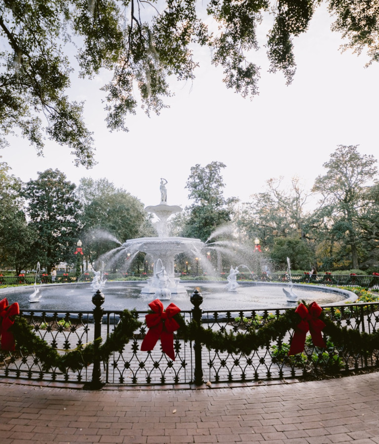 Savannah's Forsyth Park fountain surrounded by festive garlands and red bows, with holiday decorations adorning nearby lampposts beneath moss-draped oak trees.
