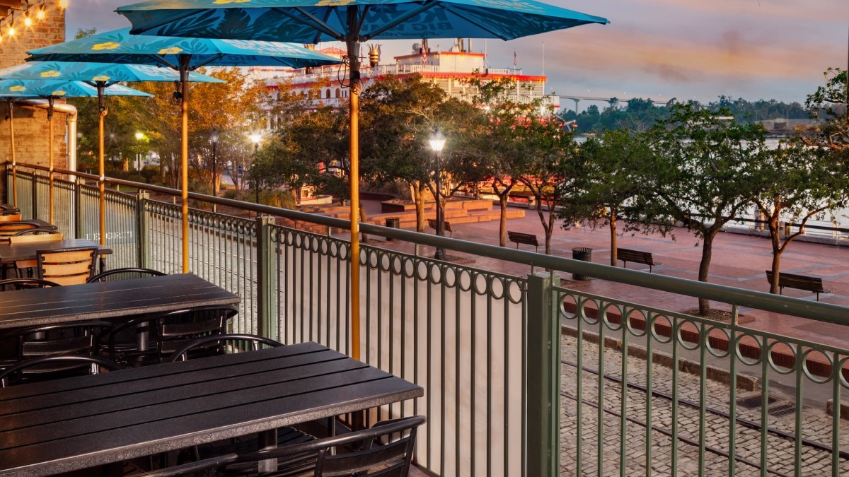 Outdoor balcony dining tables with blue umbrellas overlooking a river.