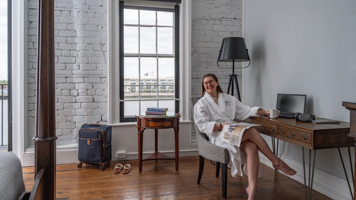 A young woman lounging in a chair in a hotel room wearing a white bathrobe.
