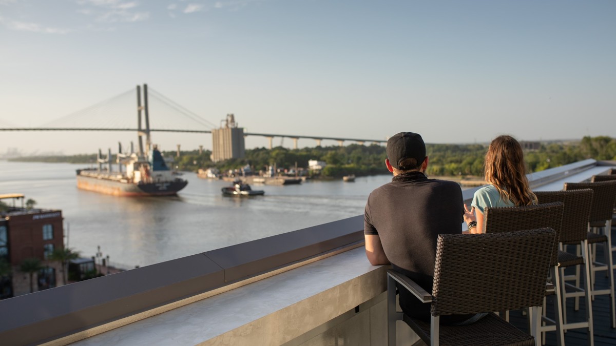A man and woman overlooking the Savannah River on a rooftop deck as ships go by.