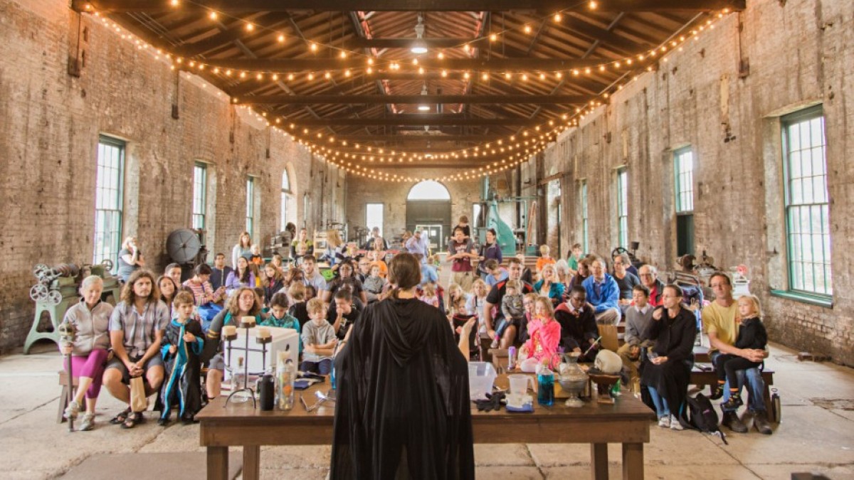 A large group of adults and children sit on benches inside a rustic brick building with string lights overhead, watching a person in a black cape leading a demonstration at a wooden table.