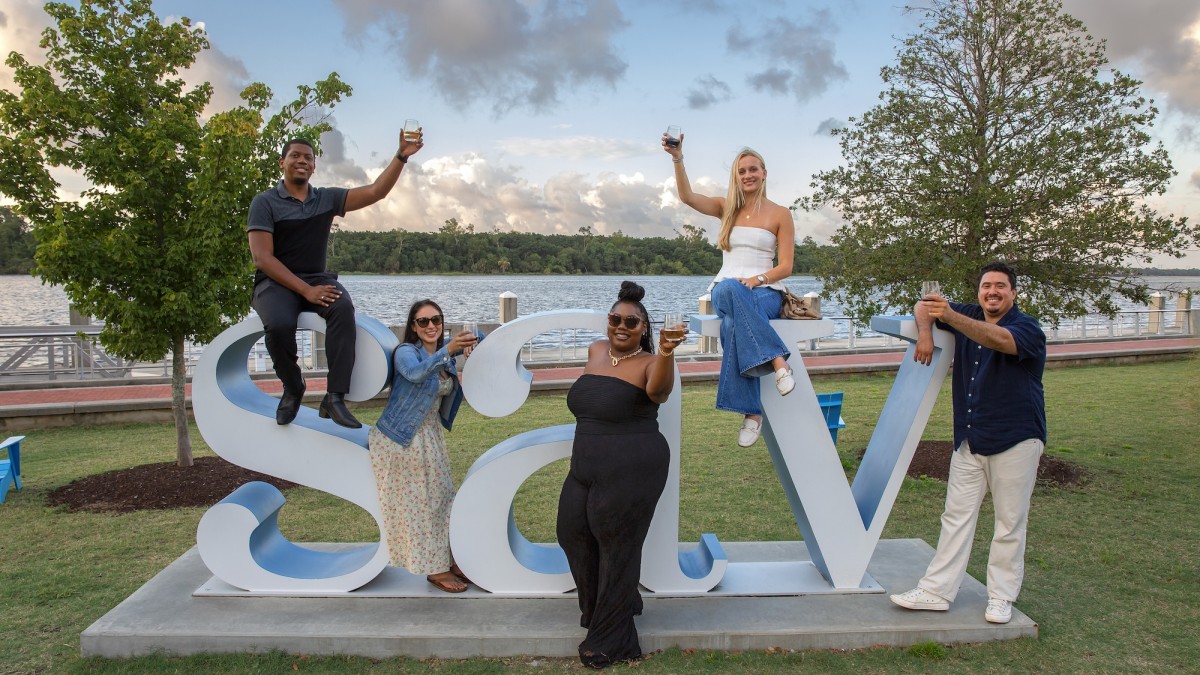 Five people raise drinks while posing on and around a large “SAV” sculpture along Savannah’s riverfront, with trees, water, and a partly cloudy sky in the background.