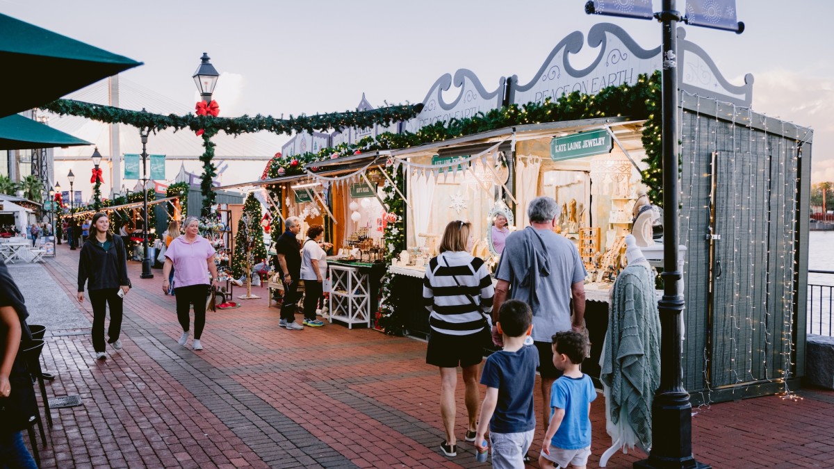 Visitors stroll through the Savannah Christmas Market, lined with festive vendor stalls decorated with garlands, bows, and twinkling lights.