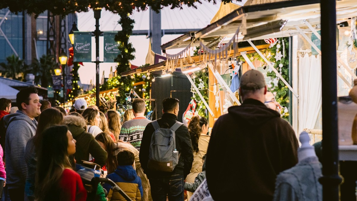 Crowds gather under garlands and twinkling lights at the Savannah Christmas Market at Plant Riverside District, exploring festive vendor stalls decorated for the holidays.