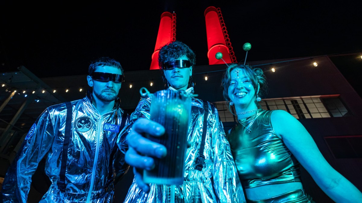 Three people dressed in futuristic space-themed costumes pose under colorful lights at the Dark Side of Electric Moon Halloween Party, with one holding a drink toward the camera and the venue’s red smokestacks glowing in the background.