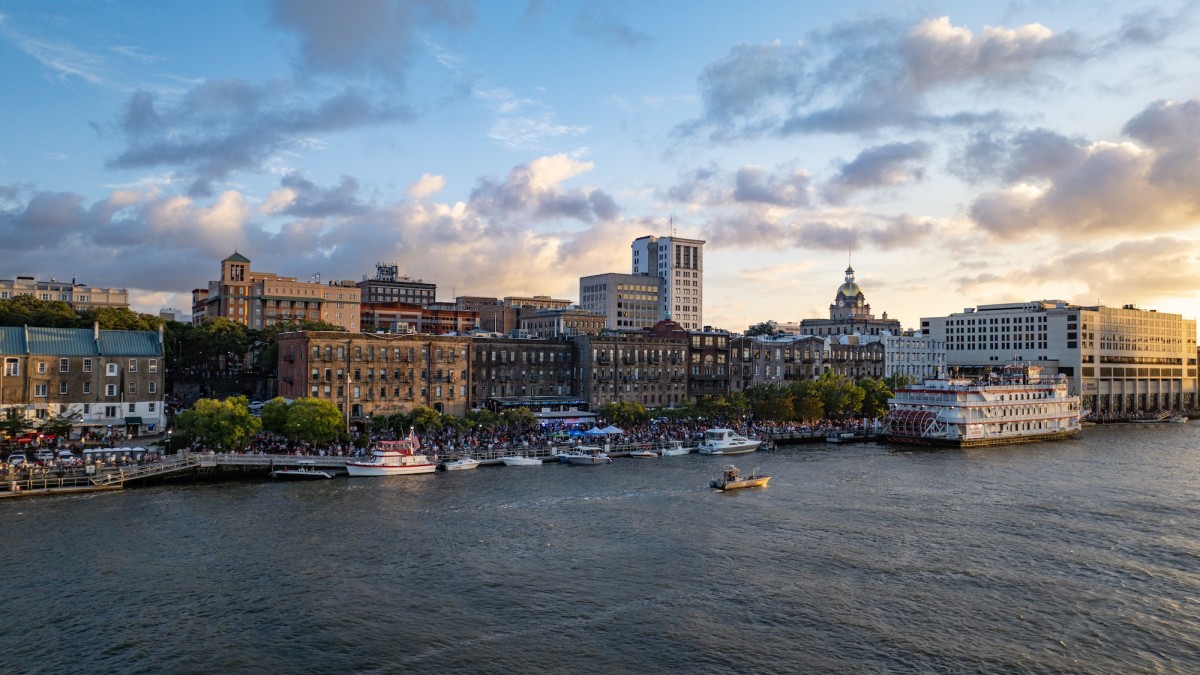 A view of Savannah’s historic River Street at sunset, with boats docked along the riverfront, crowds enjoying the waterfront, and the city skyline—including the gold-domed City Hall—glowing in the evening light.
