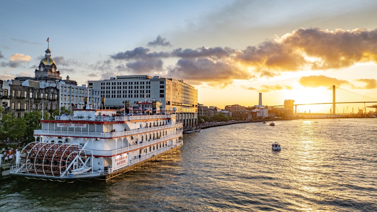 The Georgia Queen riverboat cruises along the Savannah River at sunset, with the city skyline and Talmadge Memorial Bridge glowing in the background.