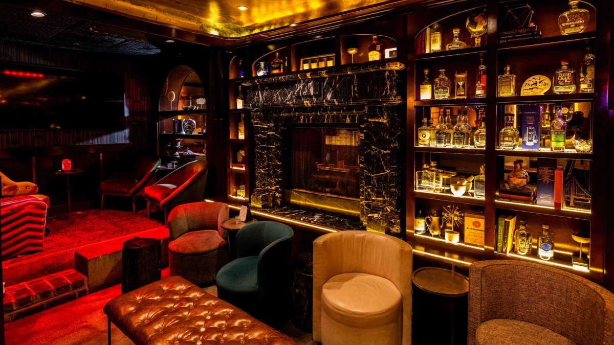 Dimly lit lounge with plush chairs, leather seating, and a black marble fireplace surrounded by backlit shelves of liquor bottles and books, creating an intimate, upscale atmosphere.