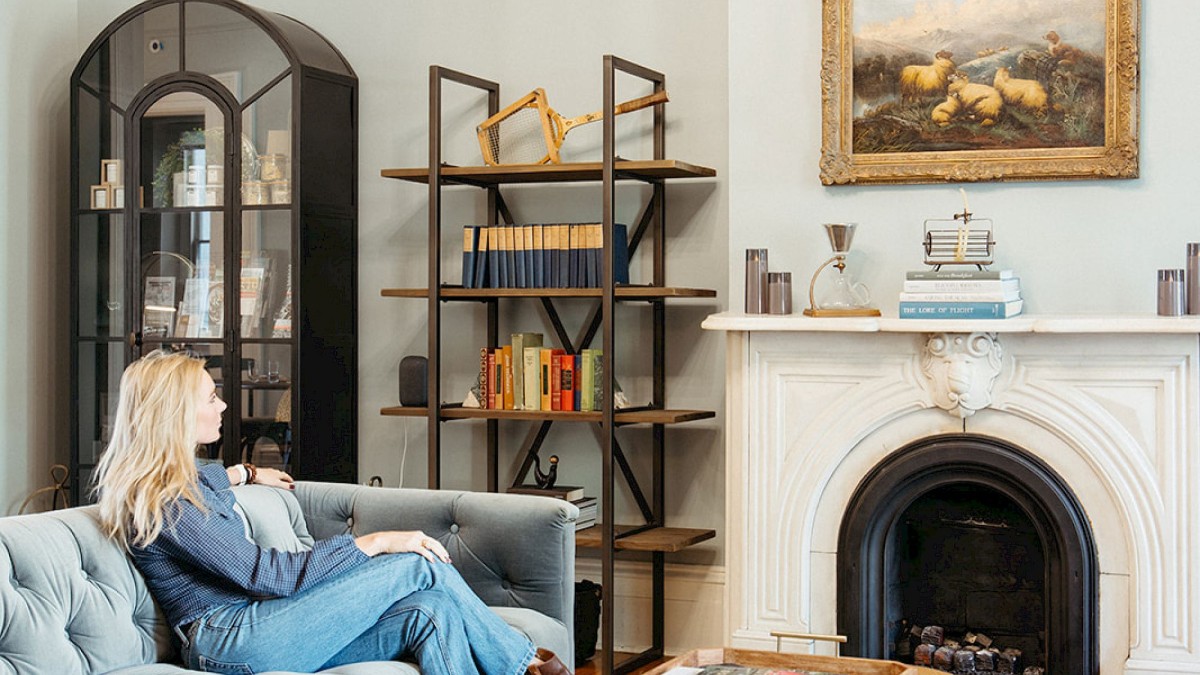 A woman relaxes on a tufted blue sofa in a bright, elegant living room at Bellwether House, featuring a fireplace, framed painting, bookshelves, and a leather ottoman.