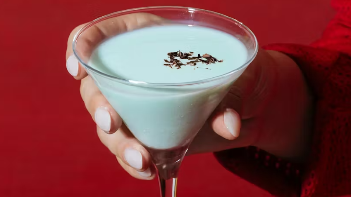 Hand holding a martini glass filled with a pale blue cocktail topped with chocolate shavings, against a red background.