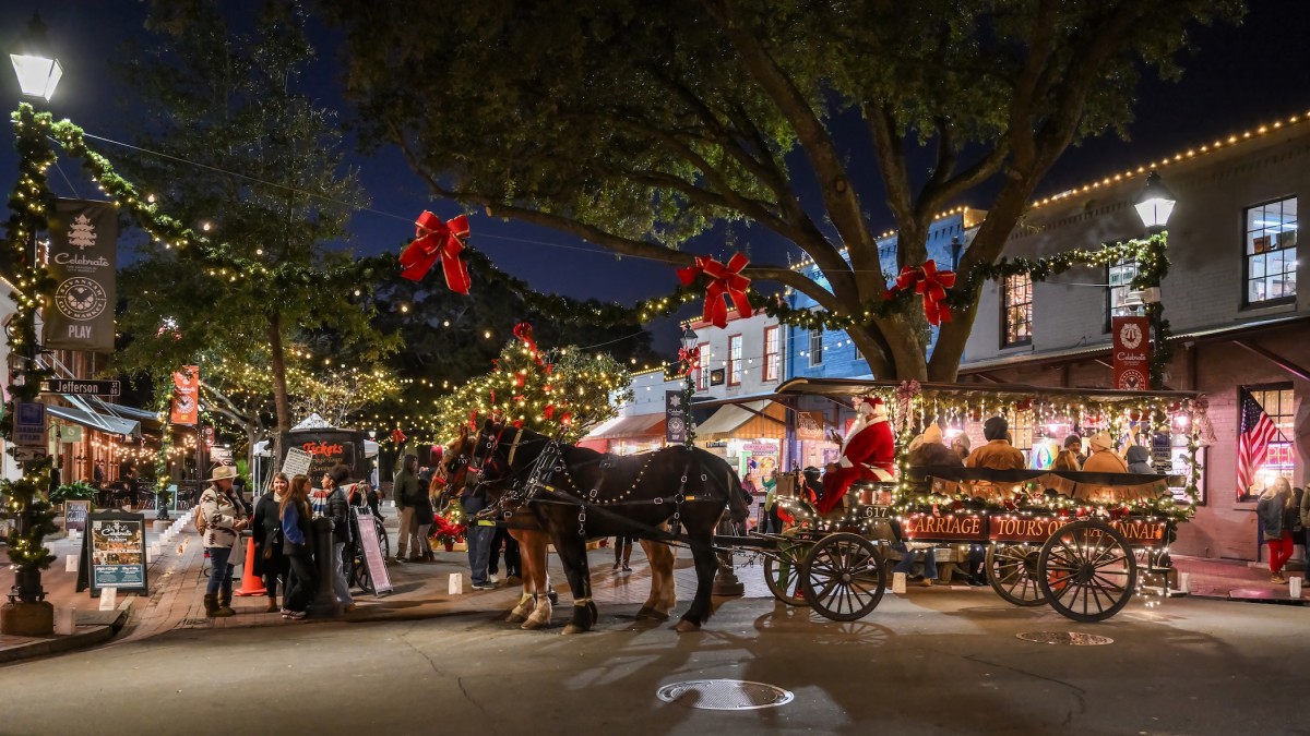 A festive nighttime scene at Savannah’s City Market during the “Celebrate the Season” holiday event. Twinkling string lights, garland, and red bows decorate the streets as visitors gather around a brightly lit horse-drawn carriage with Santa on board.