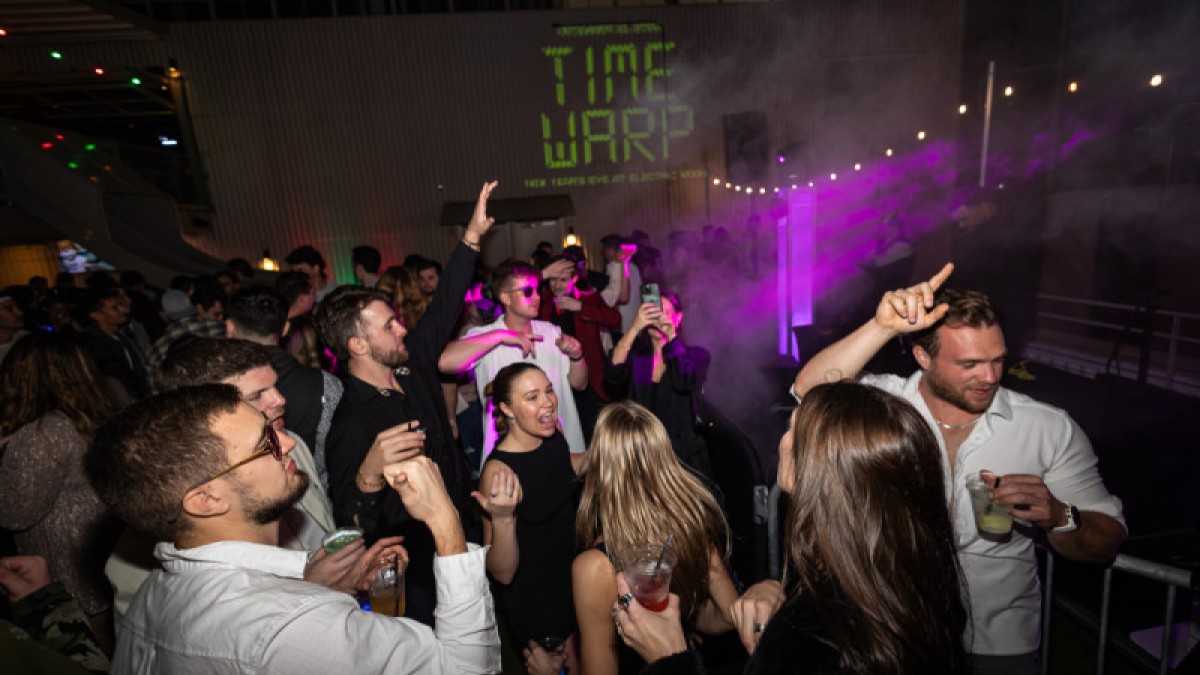 Crowd dancing and celebrating at a lively nighttime party with colorful lights, fog effects and a “Time Warp” projection in the background.