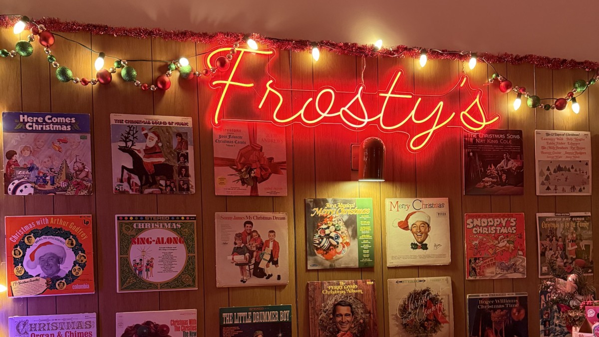 A cozy, retro holiday pop-up bar decorated with a glowing red neon sign reading “Frosty’s” above a wall of vintage Christmas album covers.