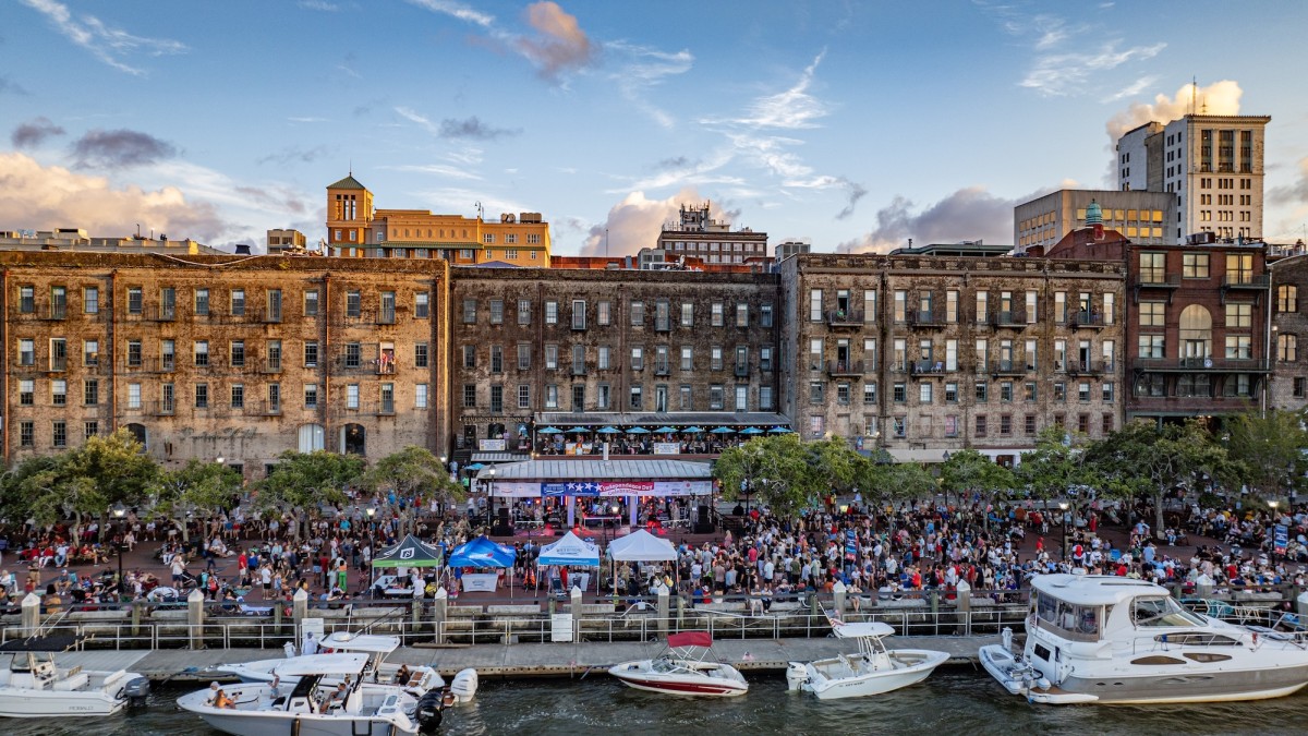 Crowds gather along Savannah’s River Street during a July 4 celebration, with live music, historic riverfront buildings and boats docked along the Savannah River.